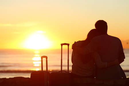 Rear view of a couple silhouette of tourists outside with suitcases on vacations enjoying watching the sun in the background on the beach at sunsetの写真素材