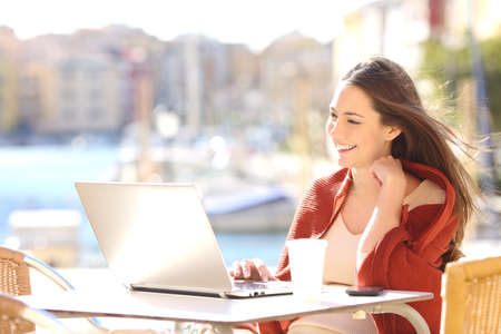 Happy casual beautiful woman watching videos or enjoying entertainment content in a laptop sitting in a coffee shop terrace outdoors in a sunny dayの写真素材