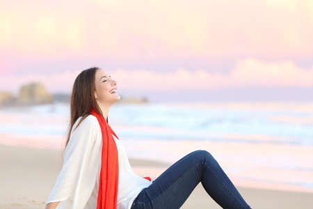 Side view of a happy woman sitting on the sand of the beach breathing fresh air at sunset with the ocean and a warmth pink sky in the backgroundの写真素材