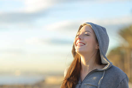 Portrait of a teenager girl wearing hood breathing deep fresh air on the beach at sunrise in a summer sunny day with a beautiful warm sky in the background