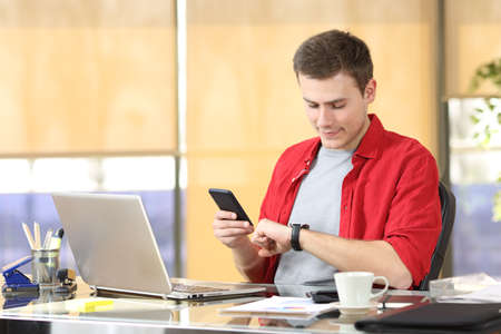 Businessman consulting a smart watch and mobile phone sitting in a desk at office