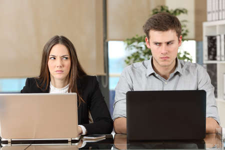 Front view of two angry businesspeople using computers disputing at workplace and looking sideways each other with envy