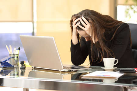 Desperate businesswoman on line worried after bankruptcy in front of a laptop with her hands in the head at officeの写真素材