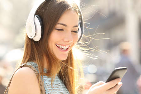 Portrait of a happy girl listening music on line with wireless headphones from a smartphone in the street in a summer sunny dayの写真素材