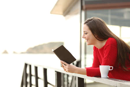 Side view of happy woman wearing a red sweater reading an ebook in an hotel or apartment on the beach on holidaysの写真素材