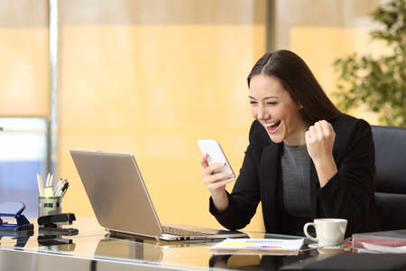 Excited businesswoman winning after achievement reading a smart phone sitting in a desktop at officeの写真素材