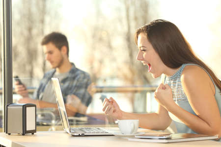 Side view portrait of an excited winner winning online consulting a laptop in a restaurant with a big window in the backgroundの写真素材