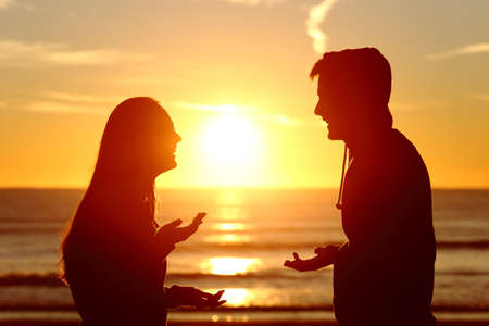 Side view of two friends or couple silhouette of teens talking happy on the beach at sunset with the sun in the middle and a warmth lightの写真素材