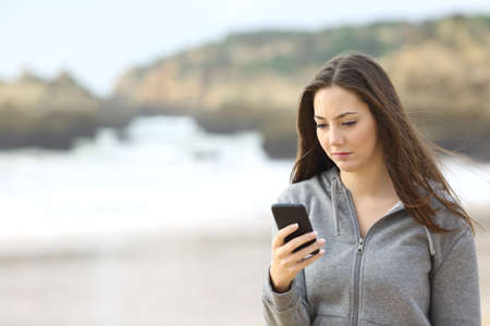 Portrait of a sad teenager walking on the beach is checking online the mobile phone waiting for a message in a rainy day