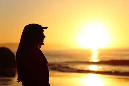 Side view of a hopeful woman silhouette looking forward with the hand on forehead at sunset with the sun horizon and sea in the backgroundの写真素材