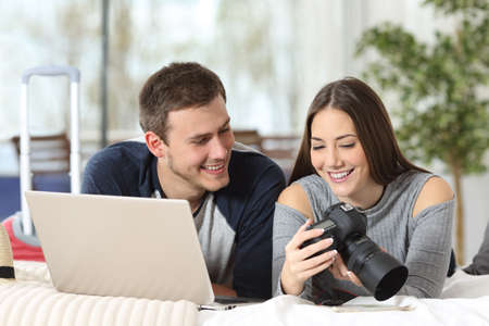 Couple of tourists watching photos from a digital camera and downloading to a laptop on the bed of an hotel room in vacationsの写真素材