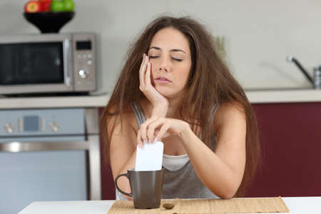 Sleepy tired girl with a bad wake up stirring coffee with the phone sitting in the kitchen at homeの写真素材