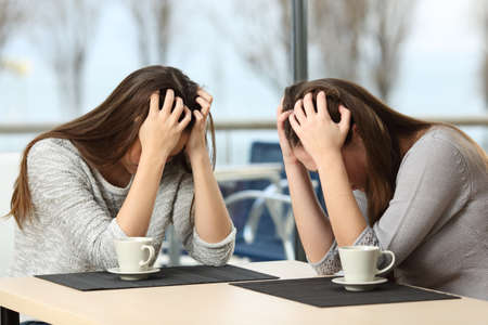 Two desperate sad girls crying with hands over head in a bar with a window with a winter backgroundの写真素材
