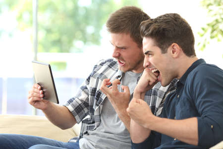 Two angry friends waiting for media content on line in a tablet with a slow connection sitting on a couch in the living room at homeの写真素材