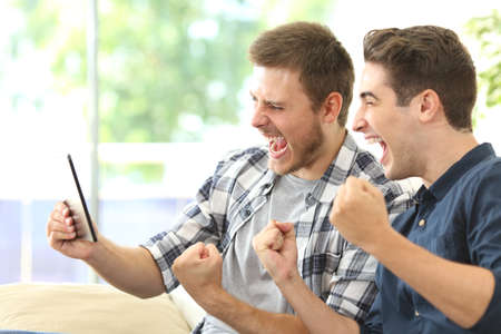 Two excited friends or roommates watching tv on line in a tablet sitting on a couch in the living room at homeの写真素材