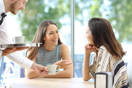 Happy friends talking looking each other sitting in the table of a bar with the waiter serving cups of coffeeの写真素材