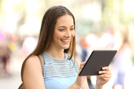 Happy woman browsing and selecting content online in a tablet walking in the street