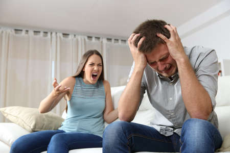 Couple arguing. Wife shouting to her desperate husband sitting on a couch in the living room at homeの写真素材