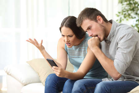 Angry couple with mobile phone sitting on a couch in the living room at homeの写真素材