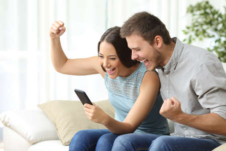 Excited couple watching media content together using a mobile phone sitting on a couch in the living room of a house