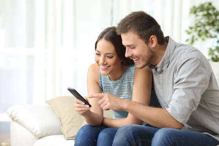 Relaxed couple or friends using a generic mobile phone together sitting on a sofa in the living room at homeの写真素材