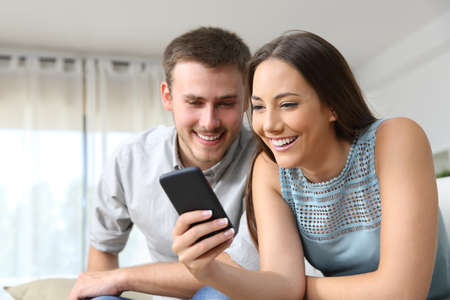 Couple consulting a mobile phone together sitting on a sofa in the living room at homeの写真素材