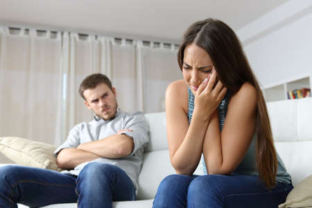 Woman lamenting domestic violence beside her angry husband sitting on a couch in the living room in a house indoorの写真素材