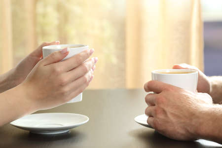 Close up of side view of a couple or friends talking with hands holding coffee cups on a table with a window and curtain in the backgroundの写真素材