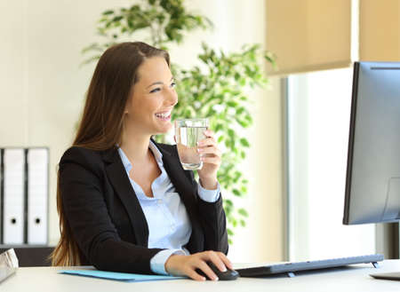 Happy businesswoman drinking water from a glass and looking through the window at officeの写真素材