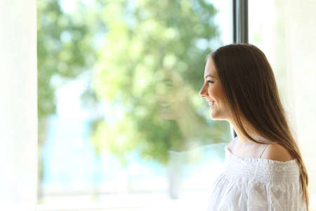 Side view of a happy lady at home looking outdoors through a window of a living room or bedroom with a sunny green backgroundの写真素材