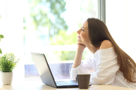 Pensive girl with a laptop daydreaming and looking through a window in a house with a green background outdoors in a beautiful dayの写真素材