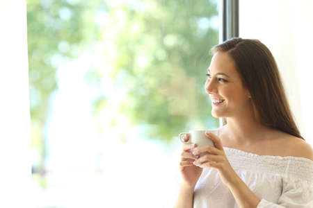 One beautiful girl tasting coffee at home and looking at horizon through a window with a green background outdoorsの写真素材