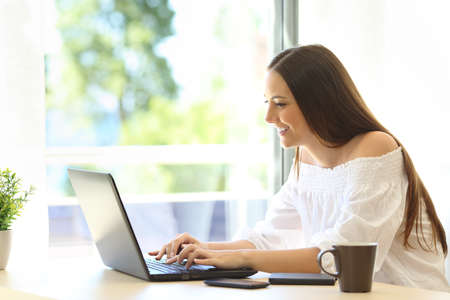 Side view of a writer writing on a laptop sitting in a desk beside a window with a green background outdoors in a warm placeの写真素材