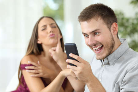 Man with a smart phone ignoring a sex proposal of his girlfriend sitting on a sofa at homeの写真素材