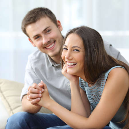 Happy couple posing and looking at you with the girlfriend in foreground sitting on a sofa in the living room at homeの写真素材