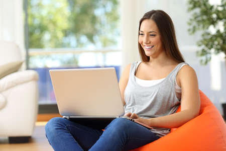 Housewife using a laptop at home sitting on an orange pouf in the living room at homeの写真素材