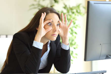 Sleepy businesswoman trying to stay awake keeping the eyes opened with the fingers watching a desktop computer monitor at officeの写真素材