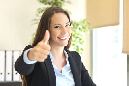 Happy businesswoman posing gesturing thumbs up and looking at camera at office with a window in the backgroundの写真素材