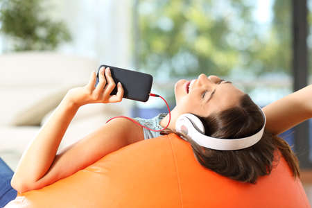 Teen listening music with headphones and smart phone lying on an orange pouf in the living room at homeの写真素材