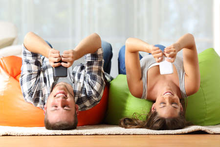 Funny roommates upside down online with their smart phones lying on orange and green poufs in the living room at homeの写真素材