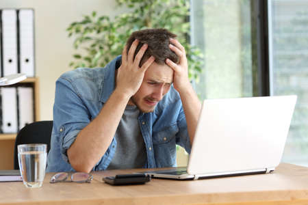 Sad entrepreneur with hands over head after reading bad news on line with a laptop at office