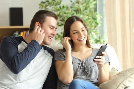 Happy couple listening music and sharing earphones sitting on a sofa in the living room at homeの写真素材