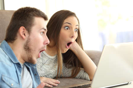 Amazed couple with open mouth on line with a laptop lying on a couch in the living room in a house...