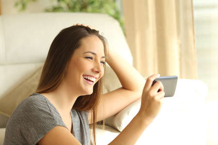 Girl watching streaming video in a smart phone sitting on the floor of the living room at home near a windowの写真素材