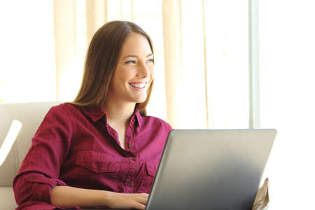 Beautiful woman on line with a laptop looking sideways through a window sitting on a couch in the living room at homeの写真素材