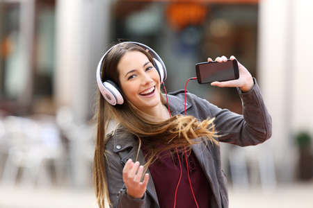 Fashion girl dancing and listening music showing a blank screen of a smart phone and looking at you in the streetの写真素材