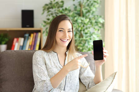 Front view of a happy woman showing a black phone screen on line and presenting a new product in a house interiorの写真素材