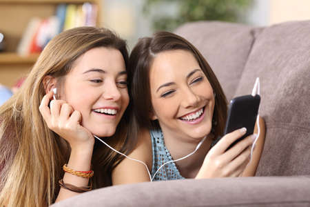 Front view portrait of two joyful friends listening music on line from a smart phone lying on a couch in the living room at homeの写真素材