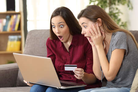 Astonished roommates buying on line with a pc and credit card sitting on a sofa in the living room at home