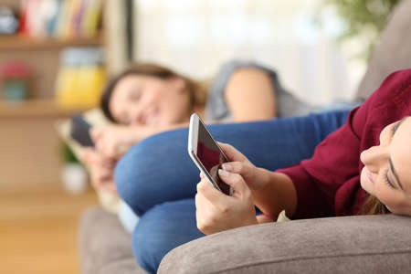 Two roommates using everyone their smart phones lying on a sofa in the living room at home with a homey backgroundの写真素材
